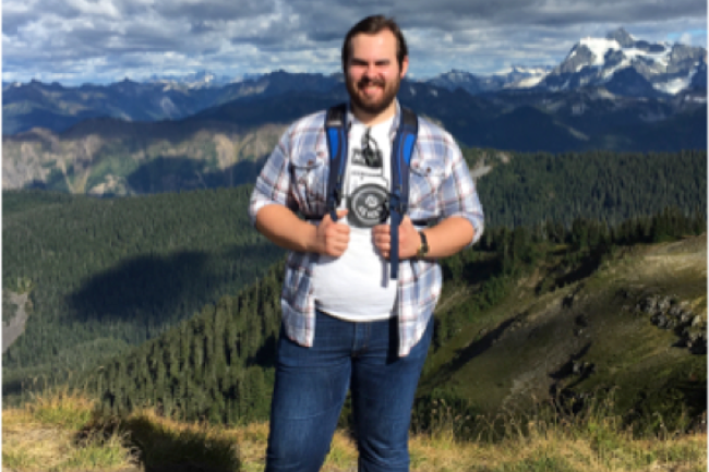 Colin stands on the top of a mountain in jeans and a white graphic shirt under an unbuttoned blue patterned button up. Behind him is a green and blue mountain range with snow only at the very peaks. He has short brown hair and a beard.