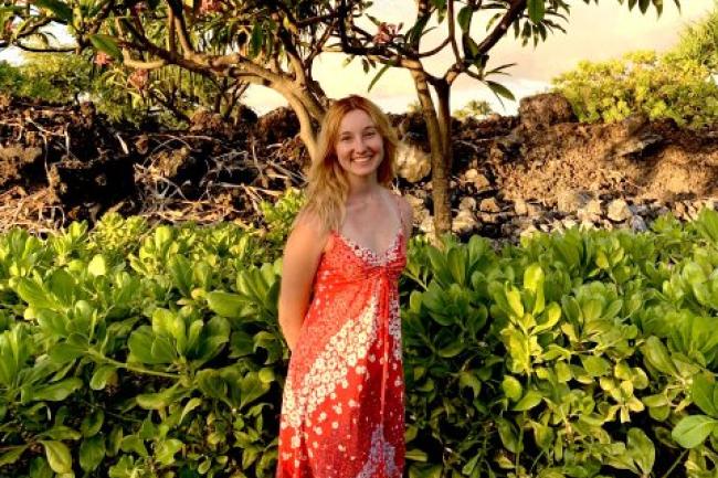 Madison has long wavy blonde hair and is wearing a loose orange, flower-patterned summer dress. She is standing and smiling in front of low, lush green foliage, a tropical tree, rocks, and a pale sky.