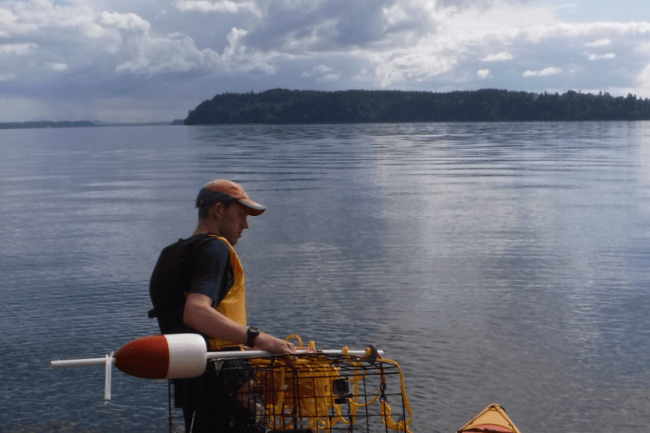 Spencer is dressed in a navy t-shirt, a yellow lifejacket vest, black shorts, black boots, and an orange and grey baseball cap. He is loading a crabbing cage and red and white buoy with yellow rope into an orange kayak. Both he and the kayak are in the shallow end of a vast expanse of water with land in the background some rowable distance away. The sky is blue with cumulous cloud formations.