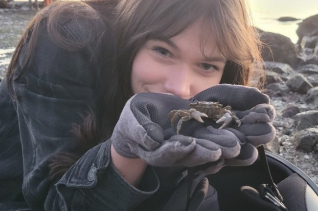 Ula has long dark hair and is wearing a black denim jacket, black leggings, and grey and black gloves. She is crouching and hiding behind her cupped hands holding a startled-looking crab. What looks like a helmet is dangling from her wrist. In the background are assorted rocks on a beach with water and a few people. 